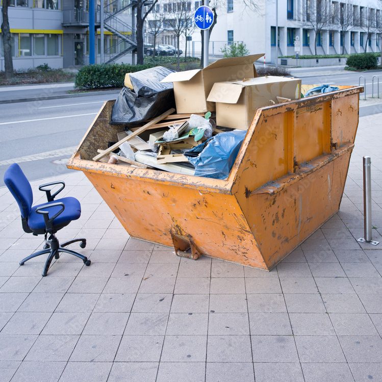 Ein gelber Abfallcontainer, überfüllt mit Müll, neben einem leeren Stuhl auf dem Gehweg.